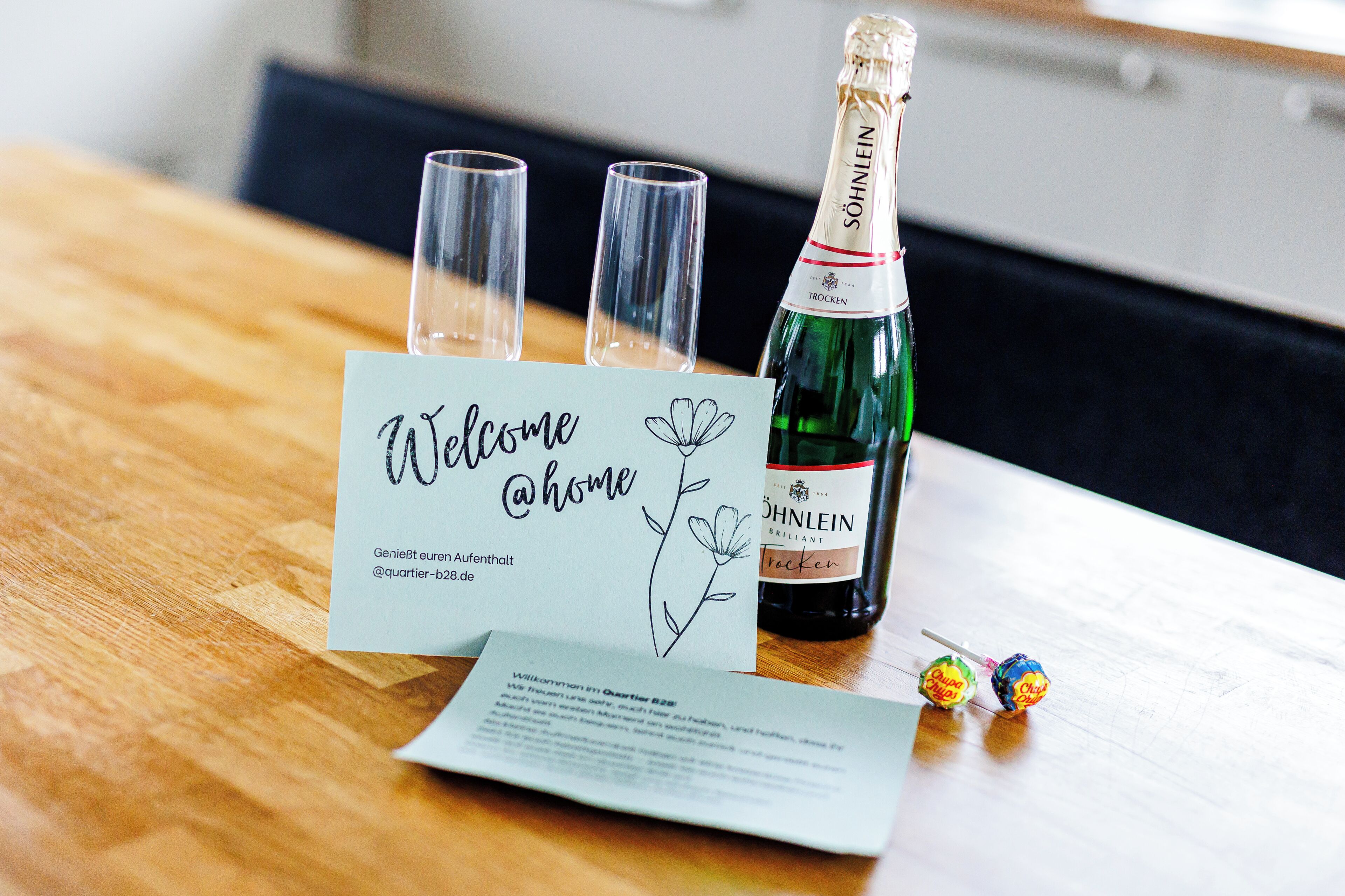 A welcome sign with a floral design, a bottle of Trocken wine, and two glasses on a wooden table.