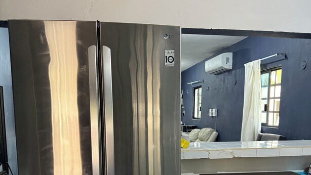 Uma geladeira de aço inoxidável com um aviso de garantia, uma mesa de madeira com um cesto, e uma bancada de cozinha com janela e cortinas.