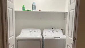 Une buanderie avec deux machines à laver et deux sèche-linge à chargement frontal, tous deux de couleur blanche, une bouteille verte et un récipient blanc posés sur l’étagère du haut.