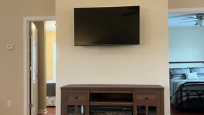 Un salon doté d’un téléviseur à écran plat fixé au-dessus d’un meuble de rangement en bois, d’une plante en pot posée sur une table basse et d’un parquet.