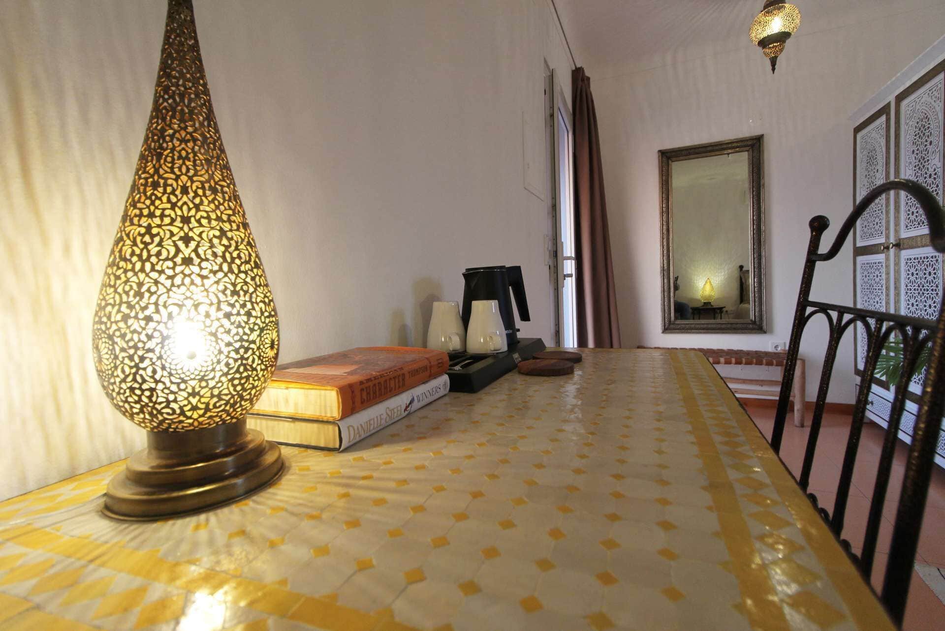 A room with a patterned floor, a table with books and a coffee maker, a lamp, and a large mirror on the wall.