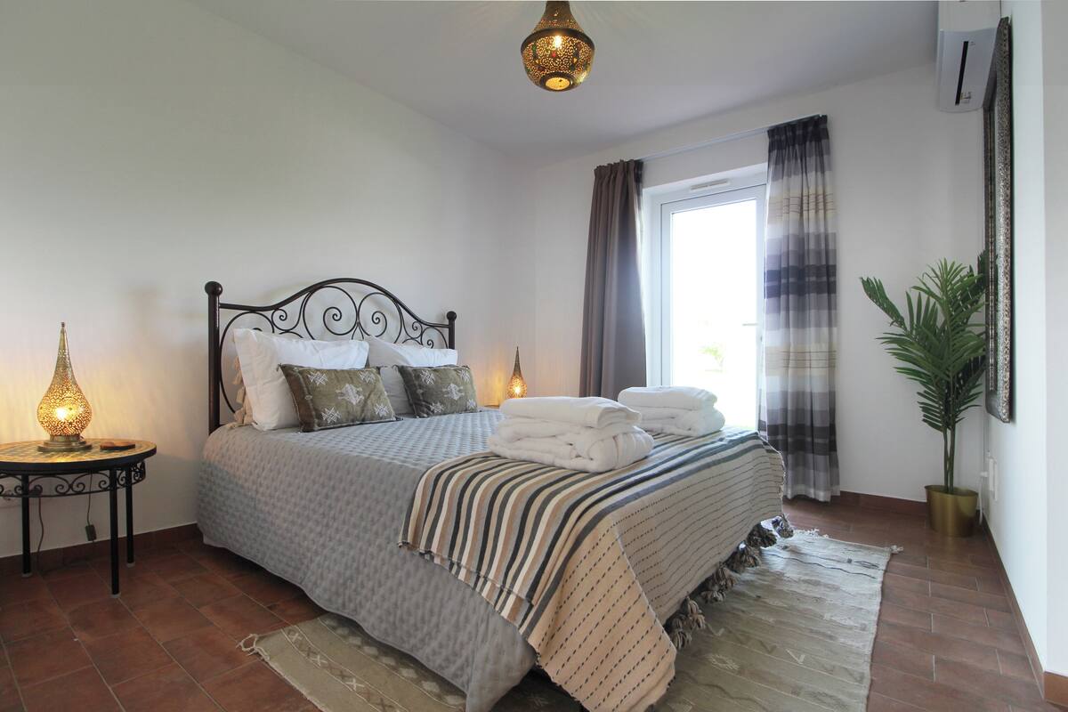A neatly made bed with a striped blanket, a bedside table with a lamp, and a potted plant.