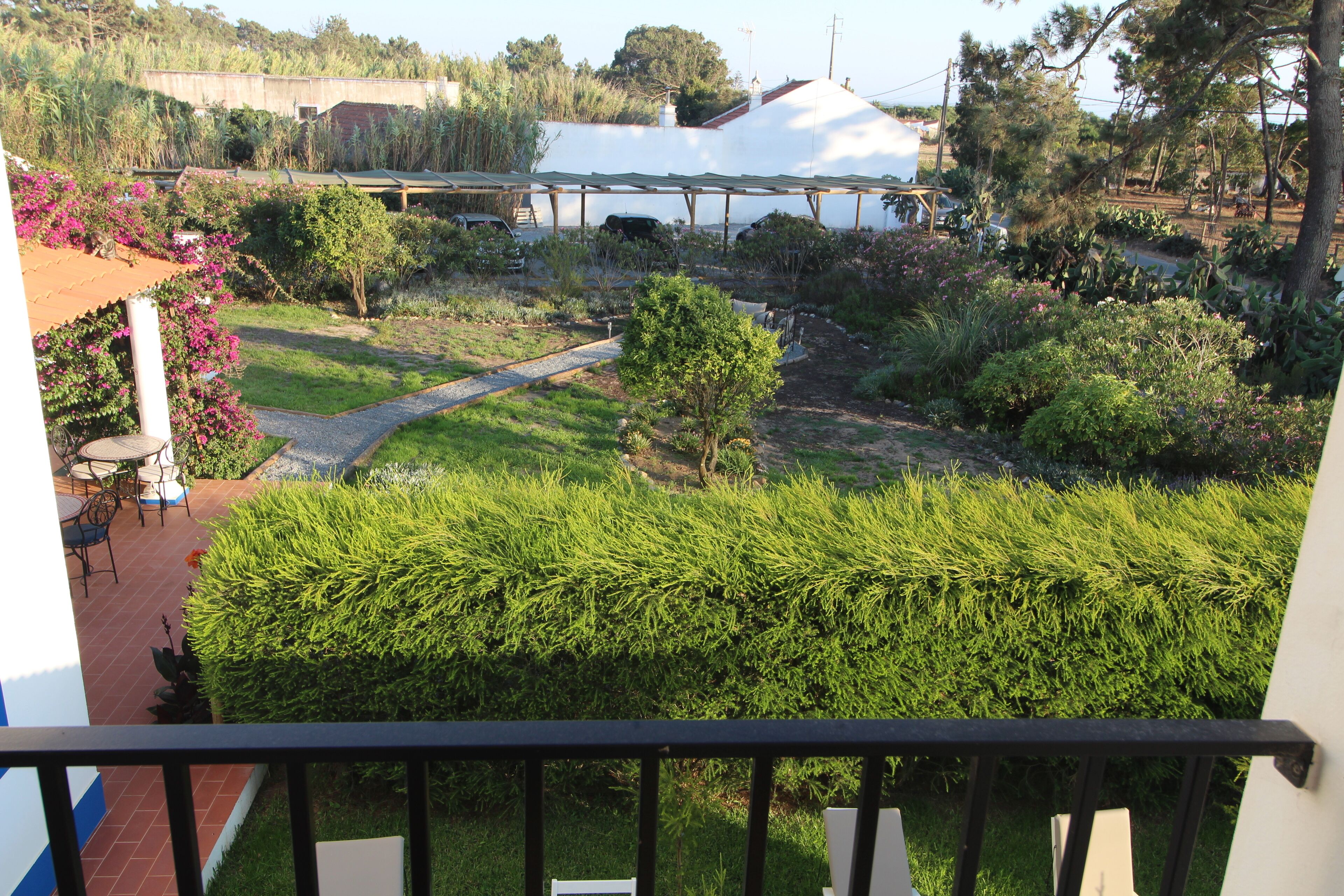 A balcony with a view of a garden, a white building, and a covered area.