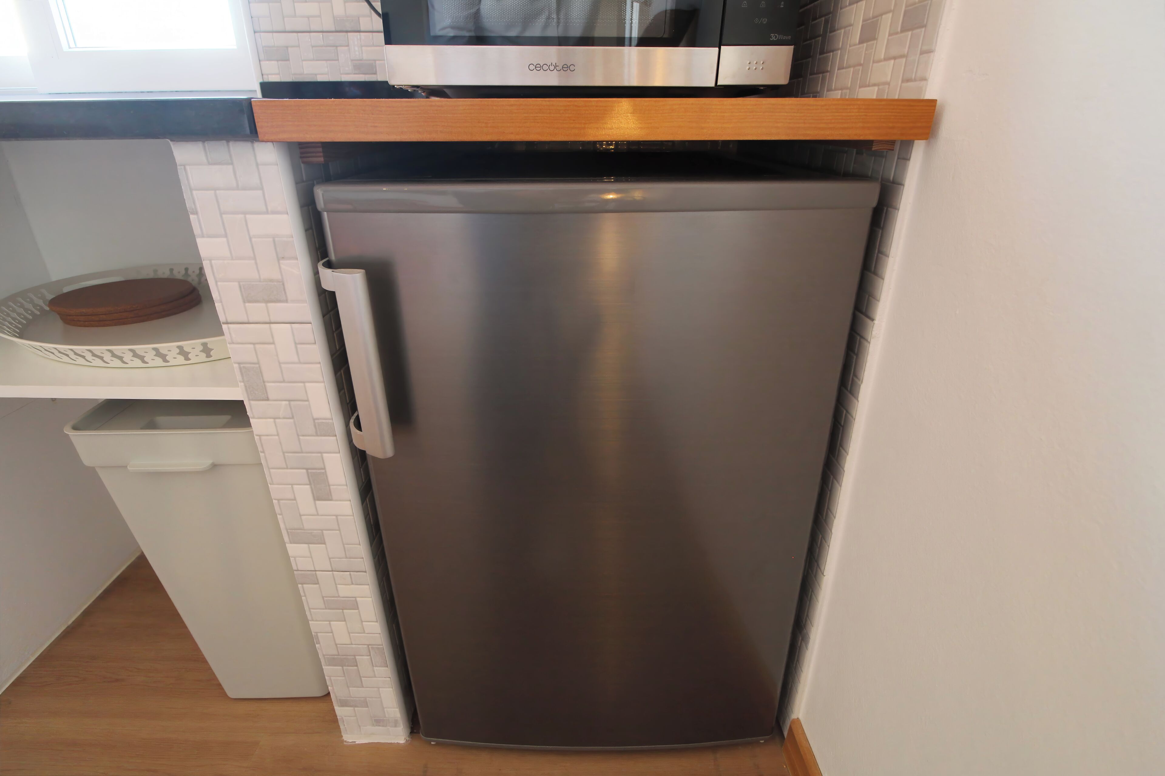 A compact kitchen with a stainless steel refrigerator, a microwave, and a wooden shelf above the fridge.