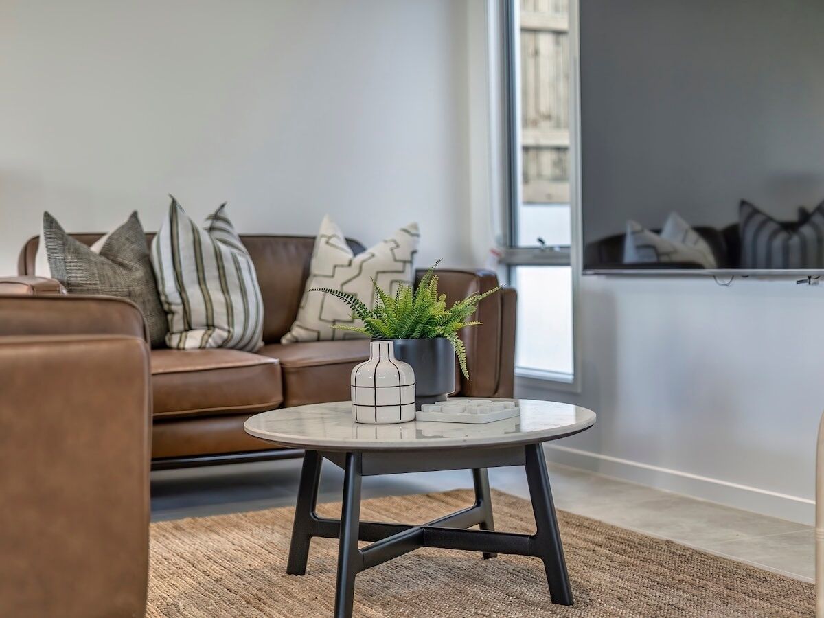 A modern living room with a brown leather sofa, a round coffee table with a white vase and green plant, and a large window providing natural light.