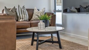 Een moderne woonkamer met een bruine leren bank, een ronde salontafel met een witte vaas en een groene plant, en een groot raam dat natuurlijk licht binnenlaat.