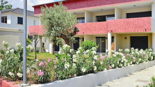 A multi-story building with a red and white facade, a central tree, and a flower-lined pathway. - Res. Marea Resort, Santa-lucia-di-moriani (Santa-Lucia-di-Moriani)