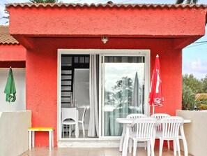Um edifício vermelho com porta de correr de vidro, cadeiras brancas, uma mesa pequena e um guarda-sol vermelho.