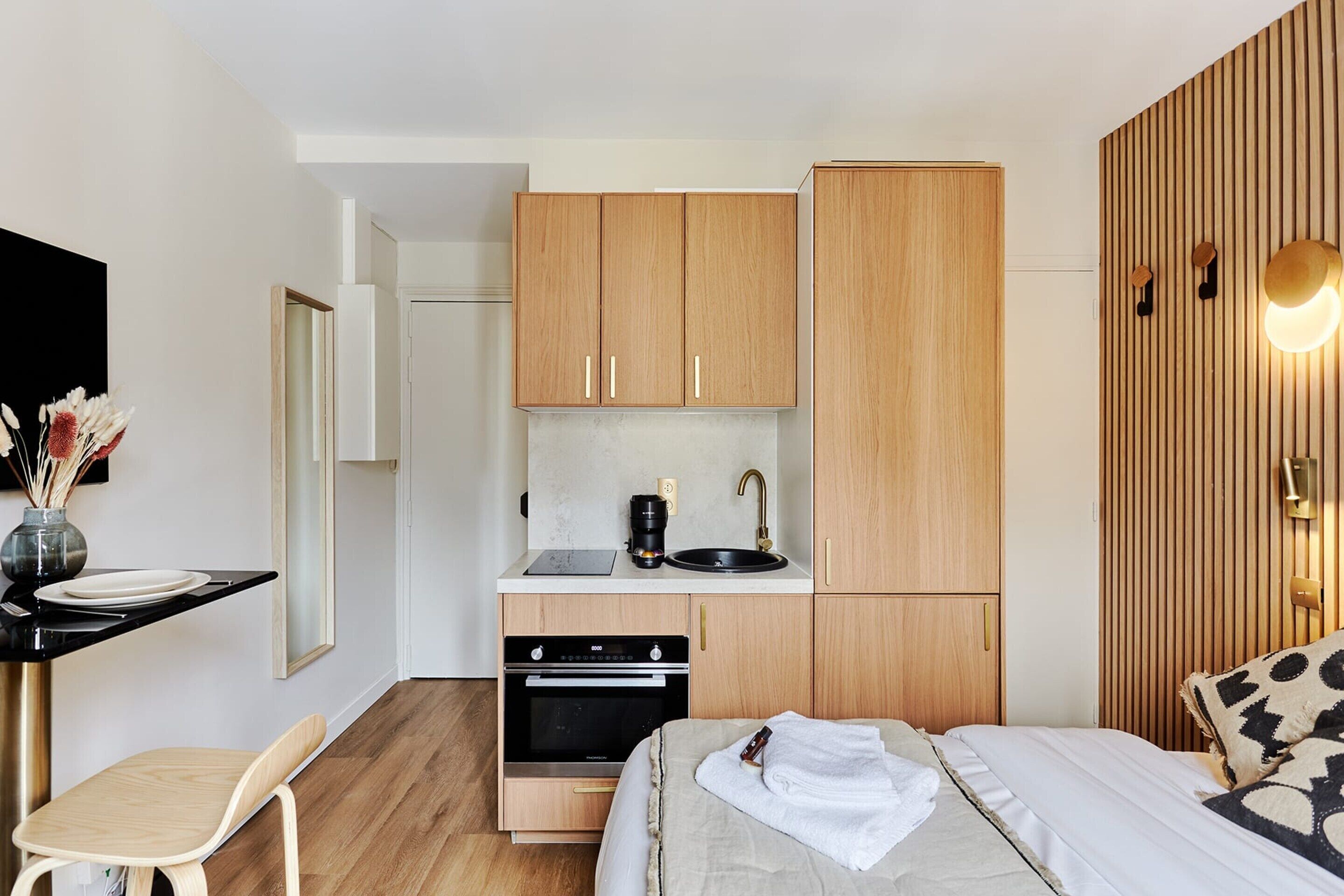 A compact bedroom with a bed, bedside table, and a small kitchenette with a sink and oven.