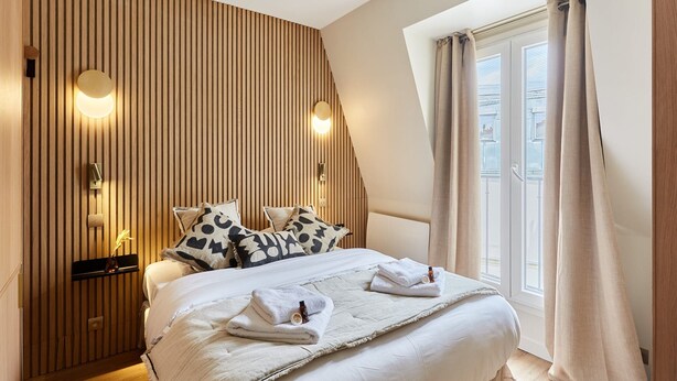 Ein modernes Hotelzimmer mit schräg angesetzter Decke, einem großen Bett mit weißer Bettwäsche, zwei Wandleuchten und einem Vorhangfenster.