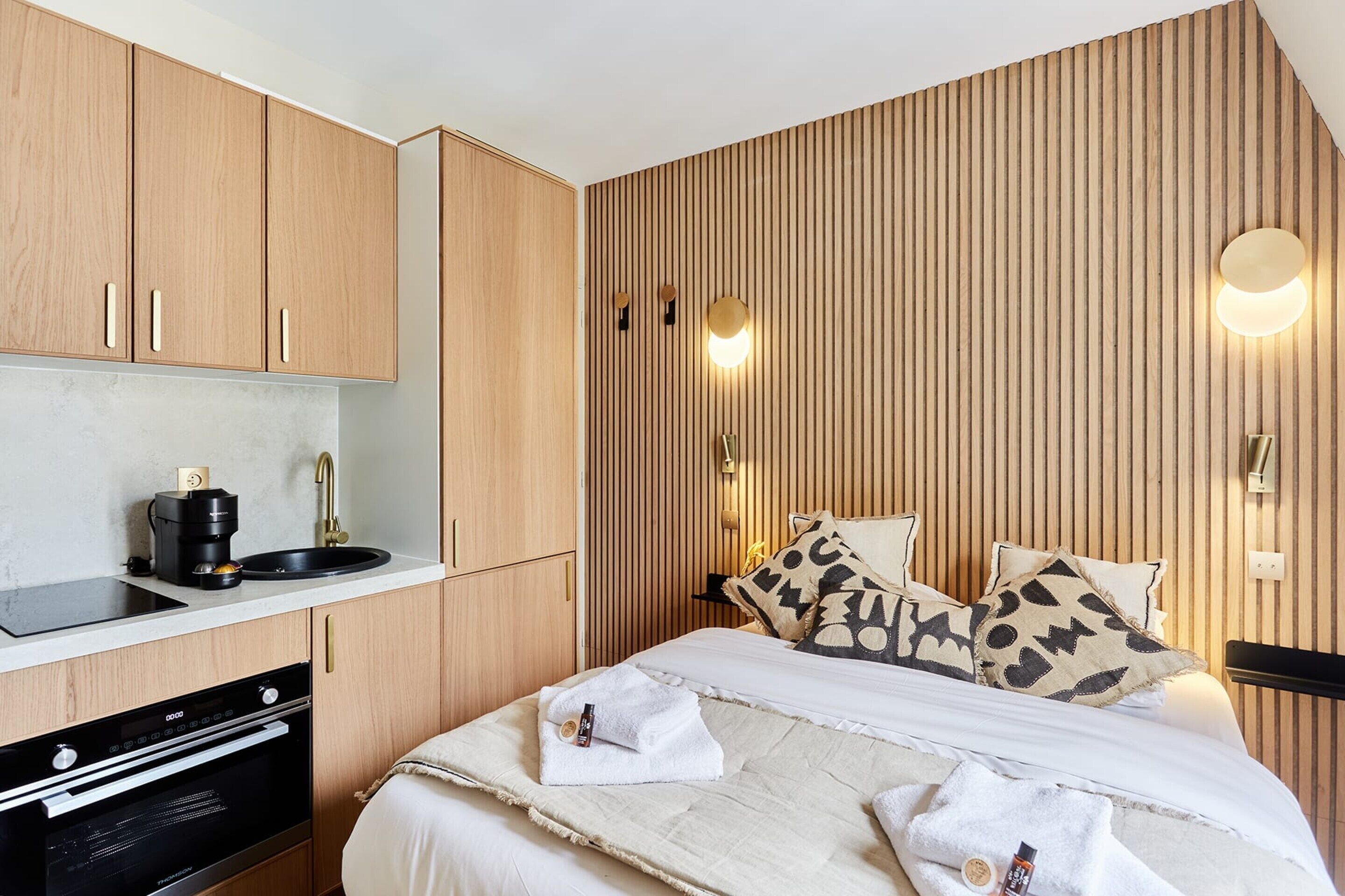 Kompakti hotellihuone, jossa on puupaneloidut seinät, valkoisilla lakanoiden peitetty sänky, pieni keittokomero, jossa on kahvinkeitin, ja seinään asennetut valaisimet.