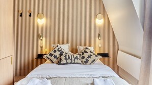 Ein Schlafzimmer mit schrägen Wänden, einer Holzvertäfelung, zwei Nachttischen mit Lampen, einem Bett mit gemusterten Kissen und einer kleinen Ablagefläche.