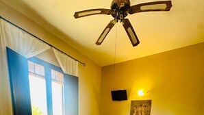 A bedroom with a bed, a desk, a chair, a ceiling fan, a window with curtains, and a wall-mounted TV.