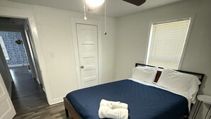 Un dormitorio con suelo de madera, una cama con colcha azul, almohadas blancas, un ventilador de techo y una ventana con persianas.