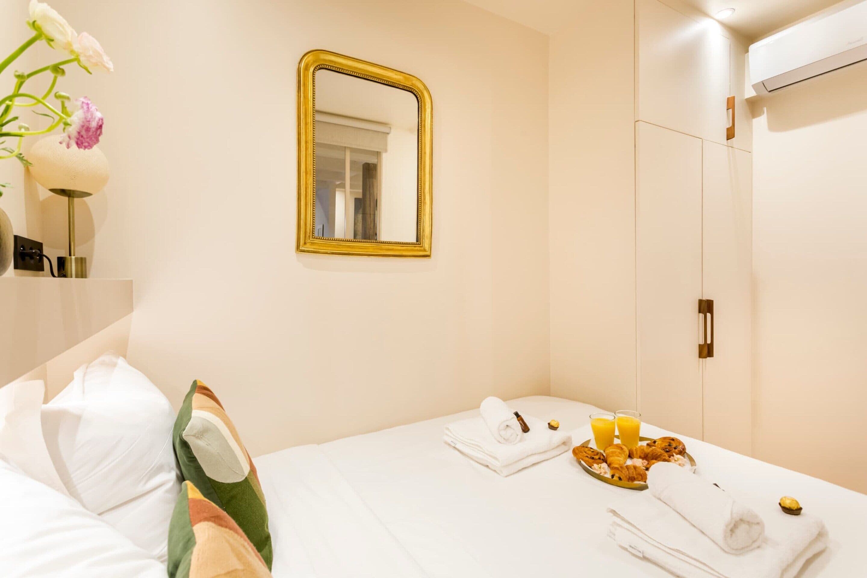 A hotel room with a bed, a mirror, a breakfast tray with croissants and orange juice, and a towel.
