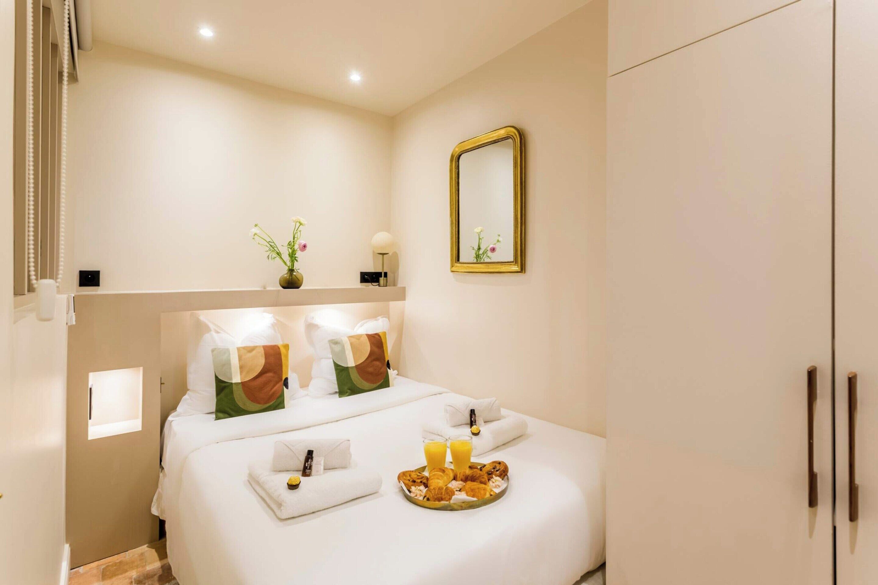 Habitación de hotel con una cama bien hecha, una mesita de noche con lámpara, un espejo y una bandeja con pasteles y jugo.