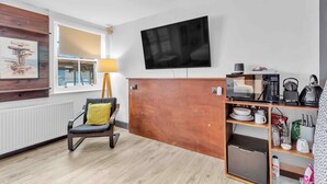 Ein modernes Hotelzimmer mit Holztresen, einem flachbildfernseher an der Wand, einer Sitzecke mit gelber Kissen und einer Küchenzeile mit Mikrowelle und Wasserkocher.