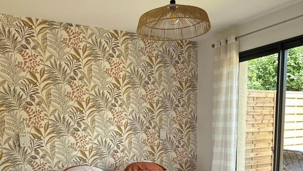 A bedroom with a patterned wallpaper, a bed with a brown bedspread, a wooden floor, and a hanging light fixture. - Le Balleré - Gîtes du Bessin (Balleroy-sur-Drôme)