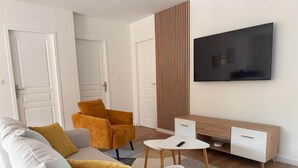 A modern living room with a sofa, armchair, coffee table, TV, and a wooden accent wall. - Le Balleré - Gîtes du Bessin (Balleroy-sur-Drôme)