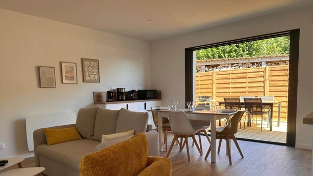 A modern living room with a sofa, a yellow armchair, and a dining area with a table and chairs. There is a kitchen area with appliances and a wooden fence visible through the sliding glass door. - Ce logement vous accueille à 25mn des plages des sites du D-DAY et de ses plages (Balleroy-sur-Drôme)