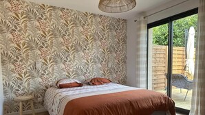 A bedroom with a large bed, a small stool, and a sliding glass door leading to a patio. - Ce logement vous accueille à 25mn des plages des sites du D-DAY et de ses plages (Balleroy-sur-Drôme)
