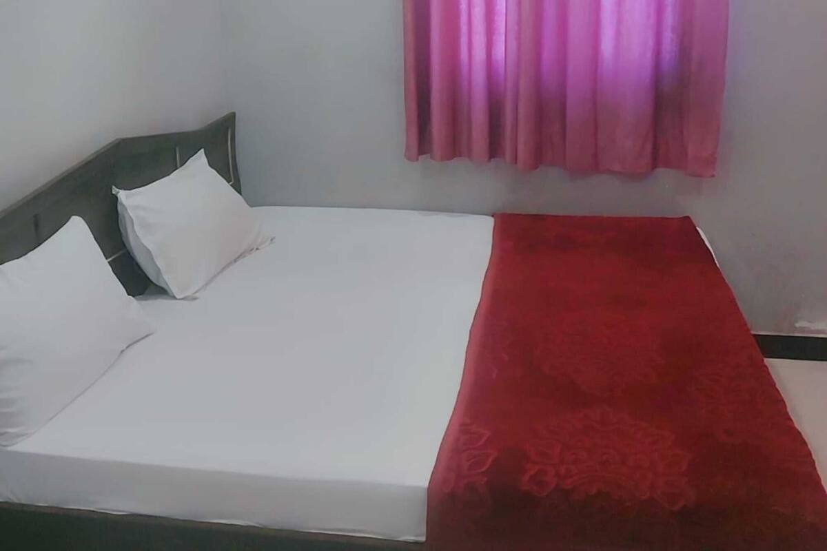 Uma cama de solteiro com roupa de cama branca e um cobertor vermelho, cabeceira cinzenta e cortina rosa.