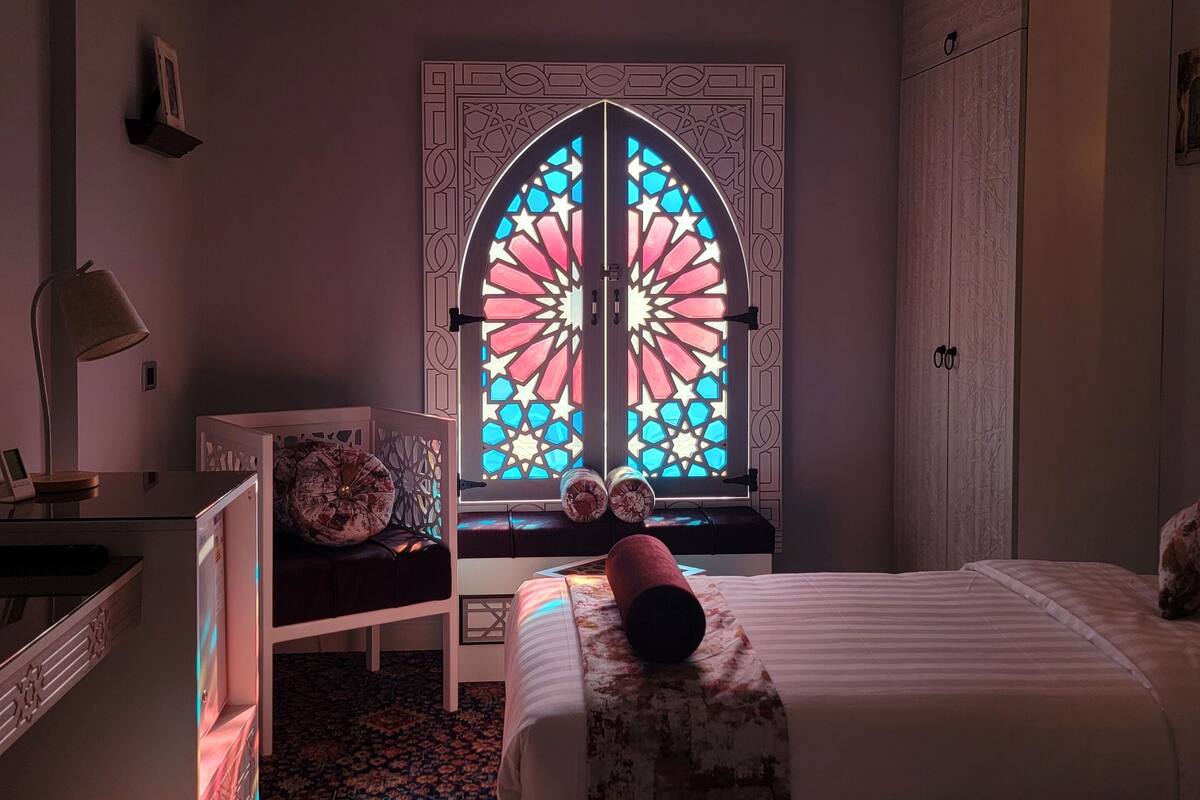 Ein Zimmer mit einem Bett, einem Schreibtisch, einem Stuhl und einem Fenster mit einem Buntglasdekor.
