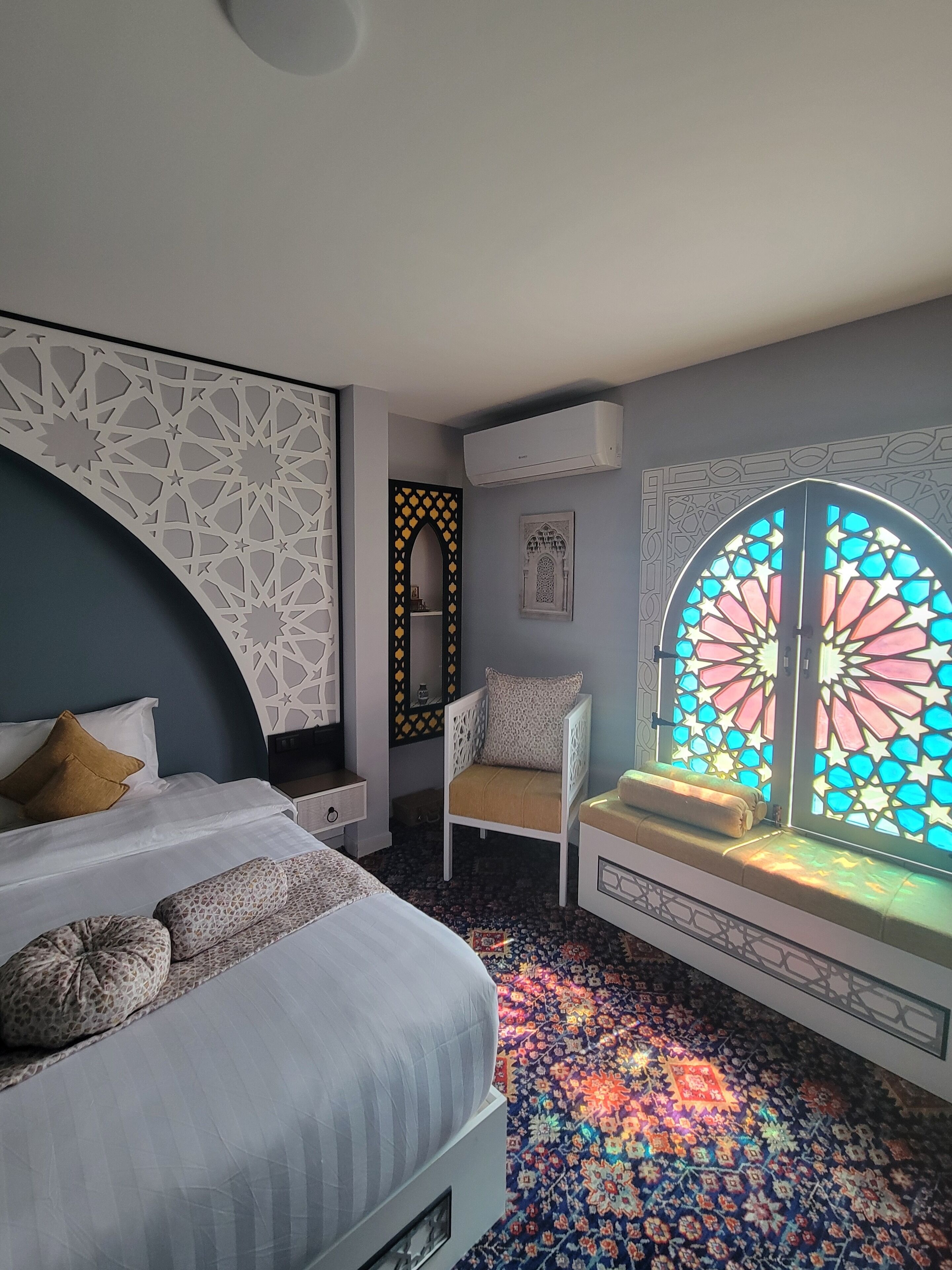 A bedroom with a bed, a chair, a window with stained glass, and a patterned carpet.