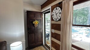 Una habitación con piso de madera, una puerta de madera oscura, una mesa redonda con una lámpara, un reloj en la pared y vista a un bosque por la ventana.
