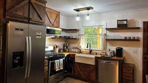 Una cocina con gabinetes de madera, un refrigerador de acero inoxidable, un microondas, una estufa y un lavavajillas. Cuenta con una ventana con cortinas, un fregadero y una cafetera.