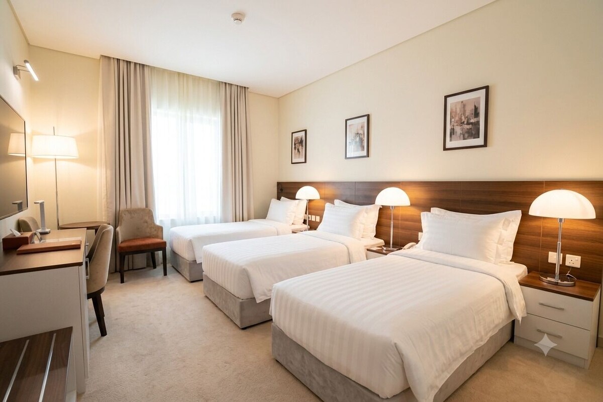 Habitación de hotel con dos camas, un escritorio, una silla y un ventanal con cortinas.