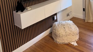 A modern living room with a flat-screen TV mounted on a white console, a fluffy pouf, and a striped wall.