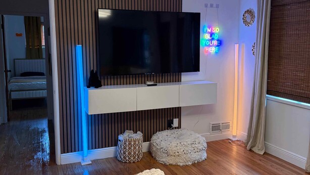 A modern living room with a flat-screen TV mounted on a white wall, wooden flooring, a brown leather sofa, and a patterned throw blanket.