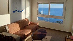 Een kamer met een groot raam en uitzicht op zee, een plafondventilator, een bank en een stoel.