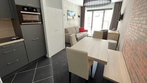 Et kompakt køkken med moderne hårde hvidevarer, et spiseområde med bord og stole, samt udsigt til det fri gennem store vinduer.