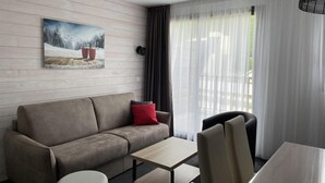Een moderne hotelkamer met een bank, een klein tafeltje en een stoel. Aan de muur hangt een schilderij en er is een raam met gordijnen.