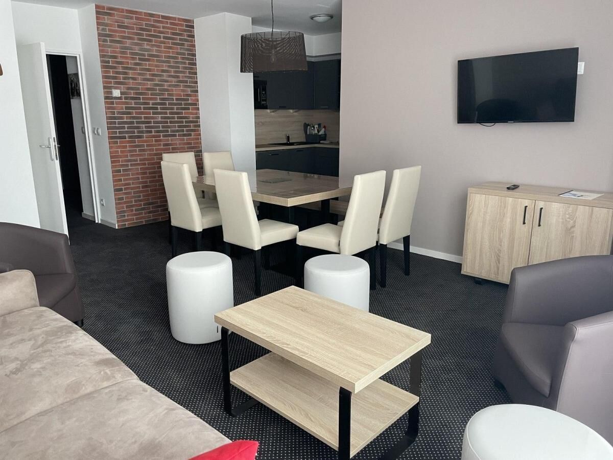 A modern living room with a dining area, a TV mounted on the wall, and a kitchen in the background.