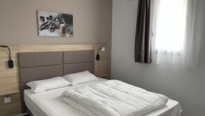 A neatly made bed with white linens, a headboard with a light wood finish, a wall-mounted artwork depicting a winter scene, and a window with sheer curtains allowing natural light.