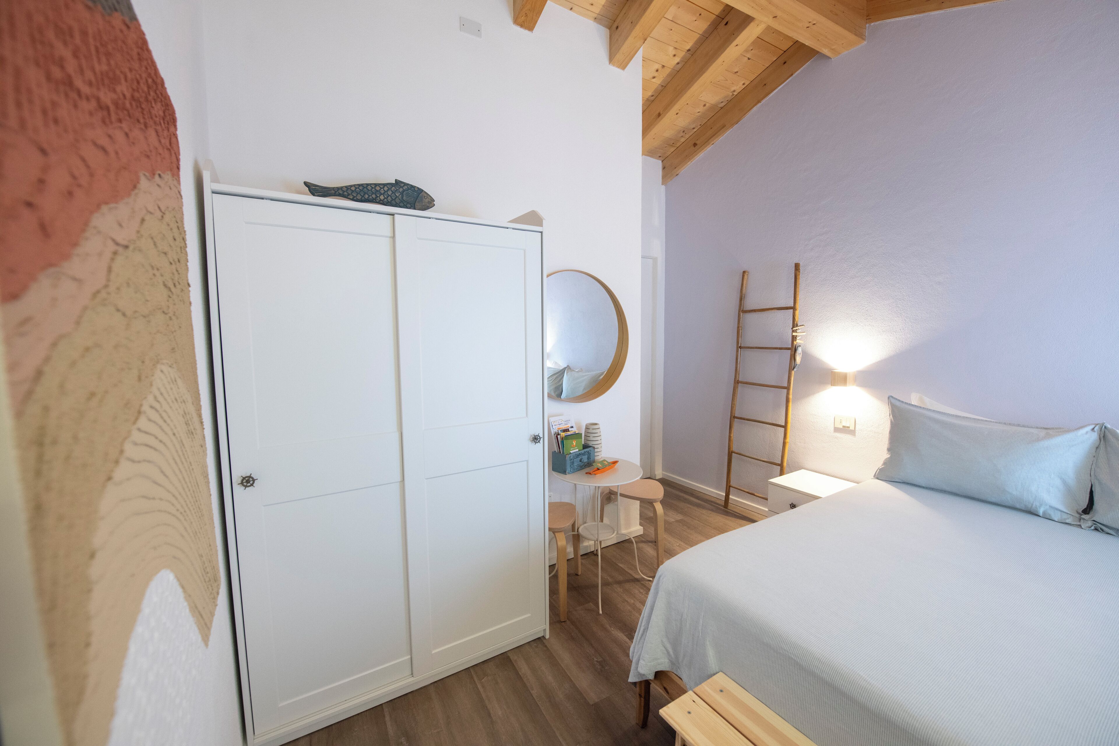 A bedroom with a bed, a wardrobe, a desk with a chair, a round mirror, and a ladder against the wall.