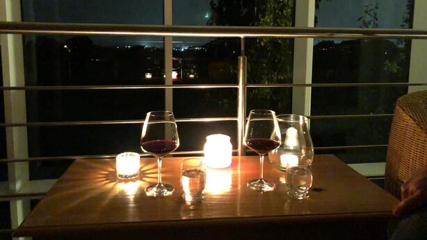 Ein Tisch mit vier Weingläsern und zwei Kerzen, vor einem Fenster mit Blick auf eine beleuchtete Stadt bei Nacht.