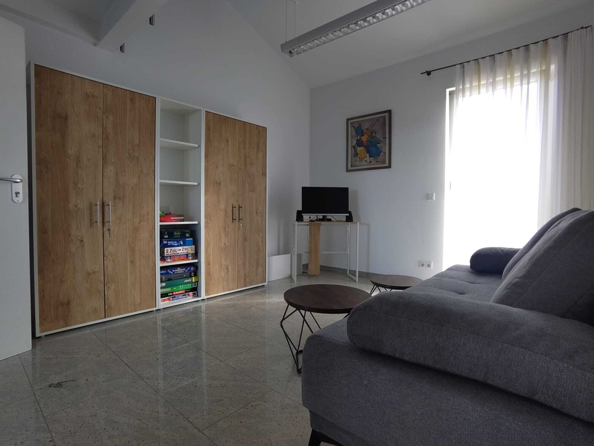 Una sala de estar con un sofá gris, un televisor sobre un mueble bajo, un armario de madera y una estantería con juegos de mesa.