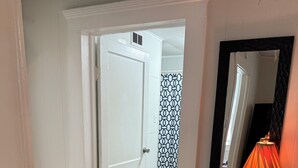 Un corridoio con pavimento in legno, una porta bianca, carta da parati decorata, uno specchio con cornice nera e un tavolo con una lampada.