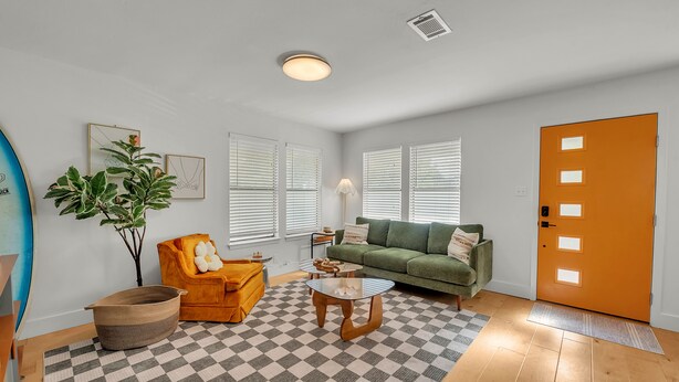 A living room with a checkered floor, a green sofa, a yellow armchair, a coffee table, a plant, and a surfboard leaning against the wall.