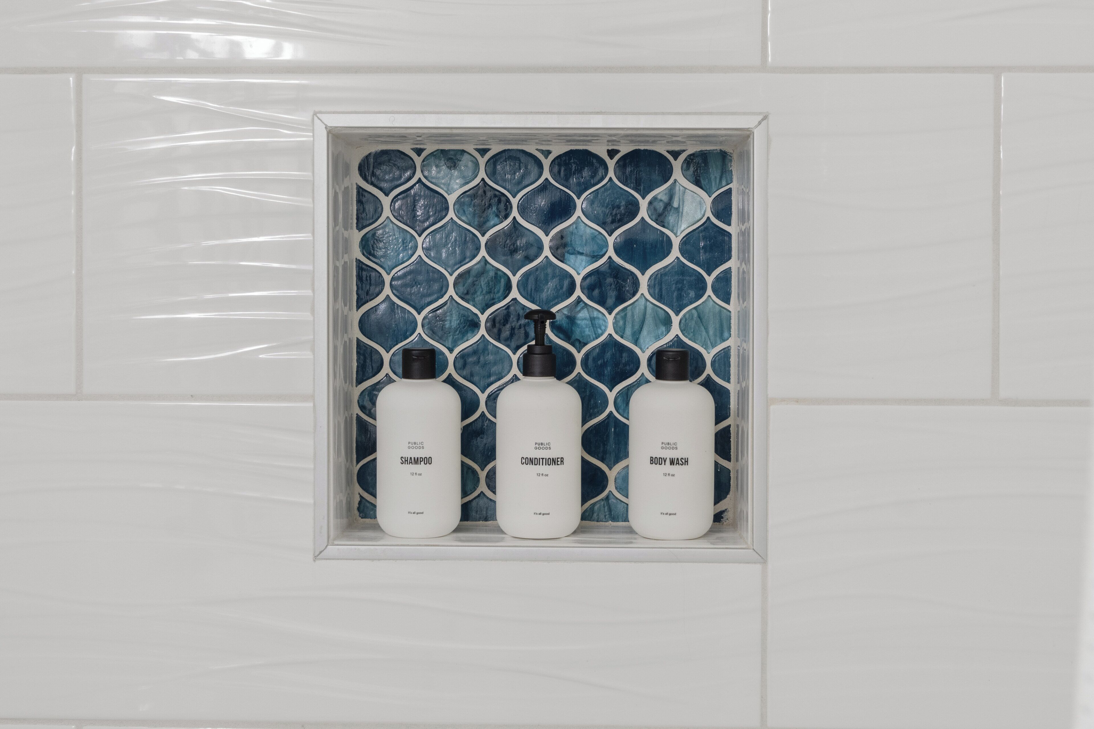 Un baño con pared de azulejos blancos y estante con tres botellas etiquetadas como 'Shampoo', 'Conditioner' y 'Body Wash'.