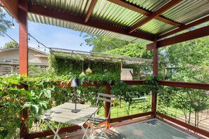 A covered patio with a table and chairs, surrounded by greenery and a view of the backyard. - Colorful Home with Private Yard with fast Wifi - Walk to East 6th - Sleeps 6 (Austin)