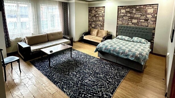 A modern bedroom with a large bed, a sofa, a small table, and a stone accent wall. - Grandi Suit Pansiyon (Elazığ)