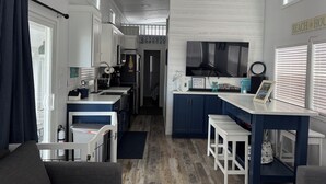 A compact kitchen with white and blue cabinetry, a large TV, and a small dining area with stools. - Delightful Tiny home with amazing views in Key Largo (Key Largo)
