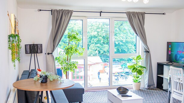 Een moderne woonkamer met een bank, een klein rond tafeltje, een witte bijzettafel en een televisie. Er zijn twee grote ramen met gordijnen, een potplant en een hangplant aan de muur.