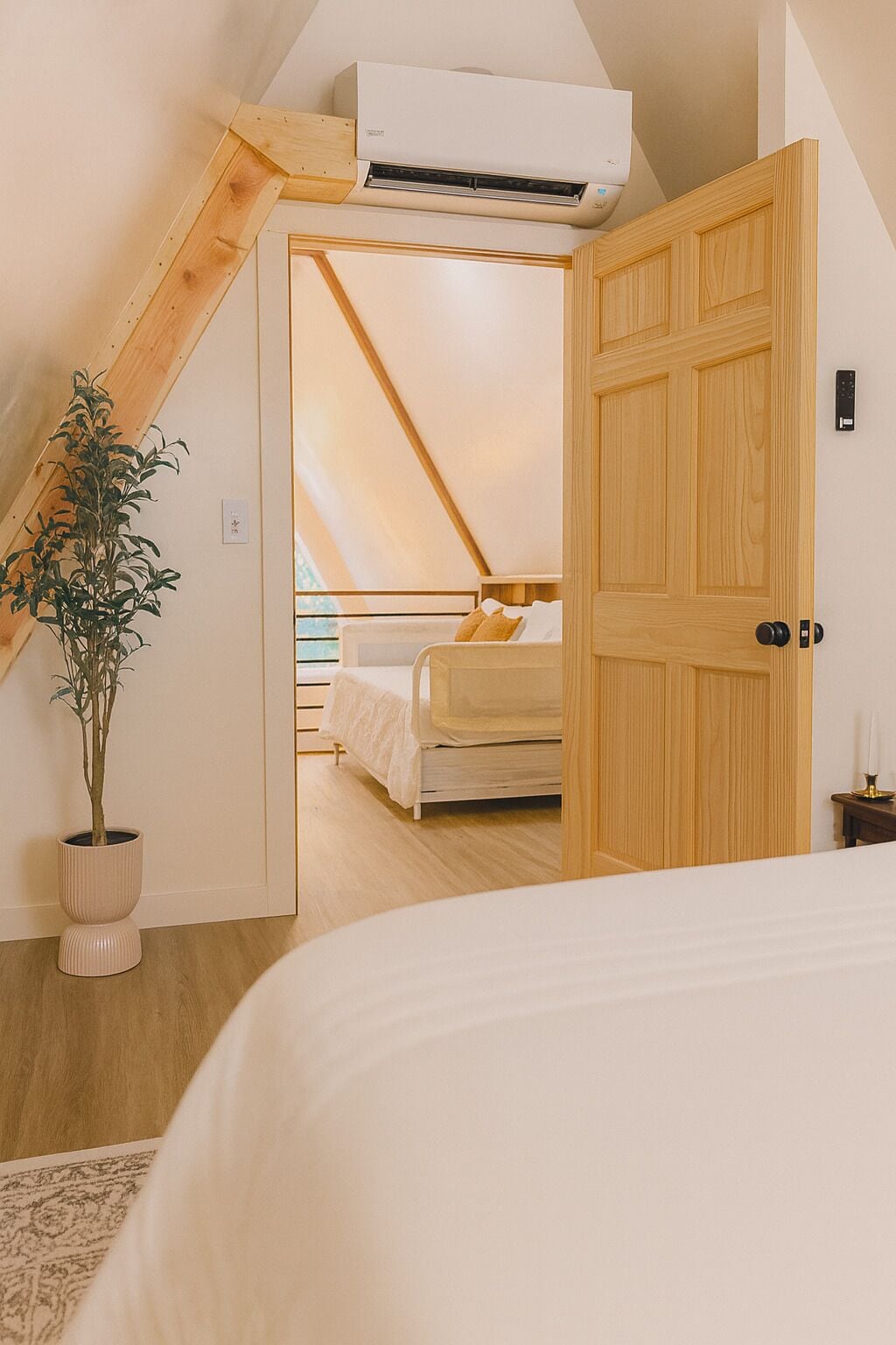 A bedroom with a sloped ceiling, a wooden door, a potted plant, and a bed with white bedding.