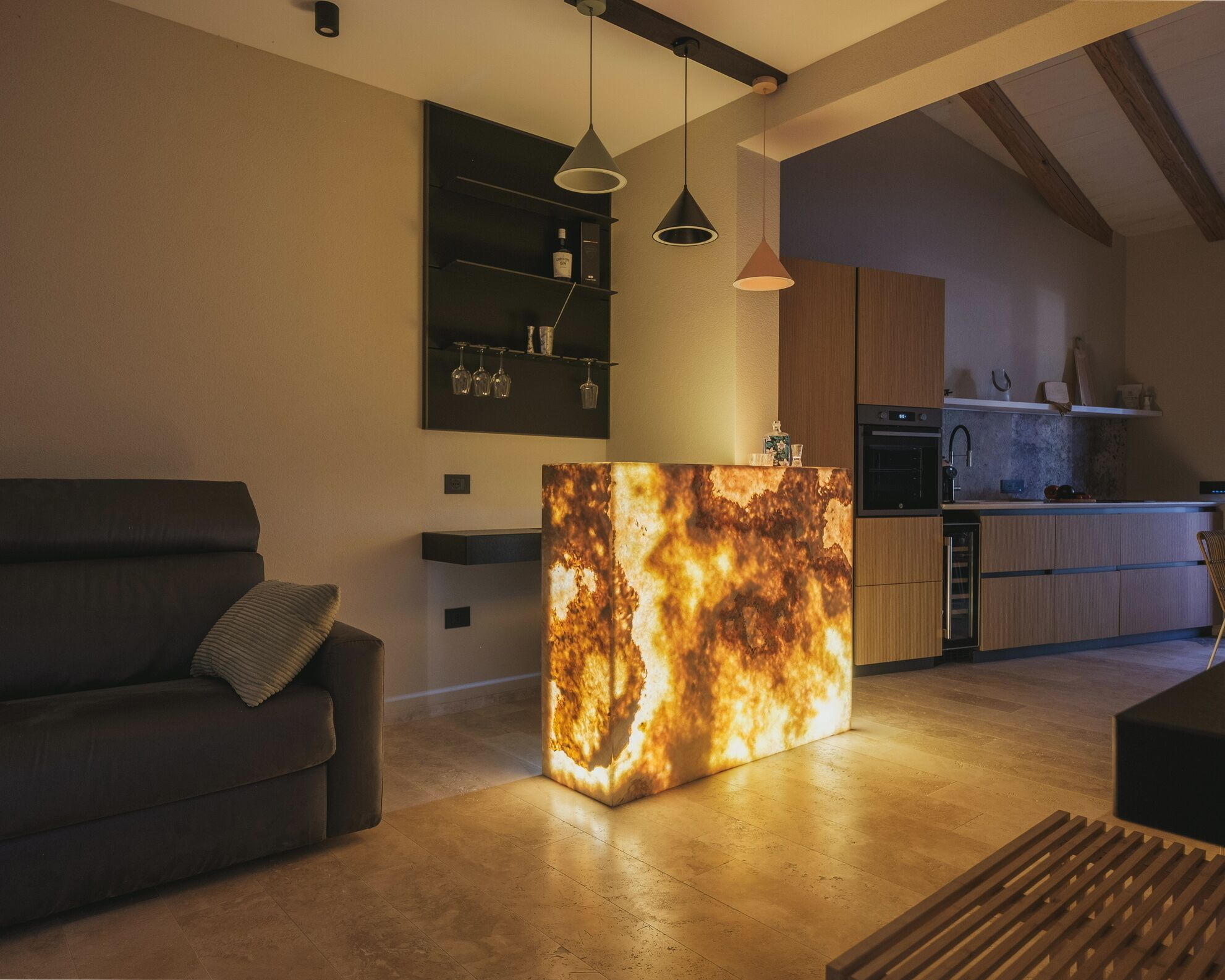 Un salon moderne avec un canapé foncé, une table basse originale à la texture particulière, des luminaires suspendus et un coin cuisine doté de meubles en bois et d’un four encastré.